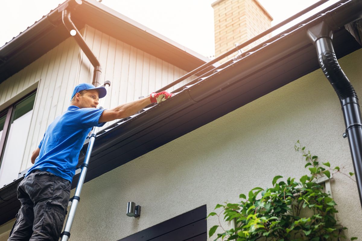 A man on a ladder meticulously cleaning the roof of a house, showcasing professional roofing service in action.